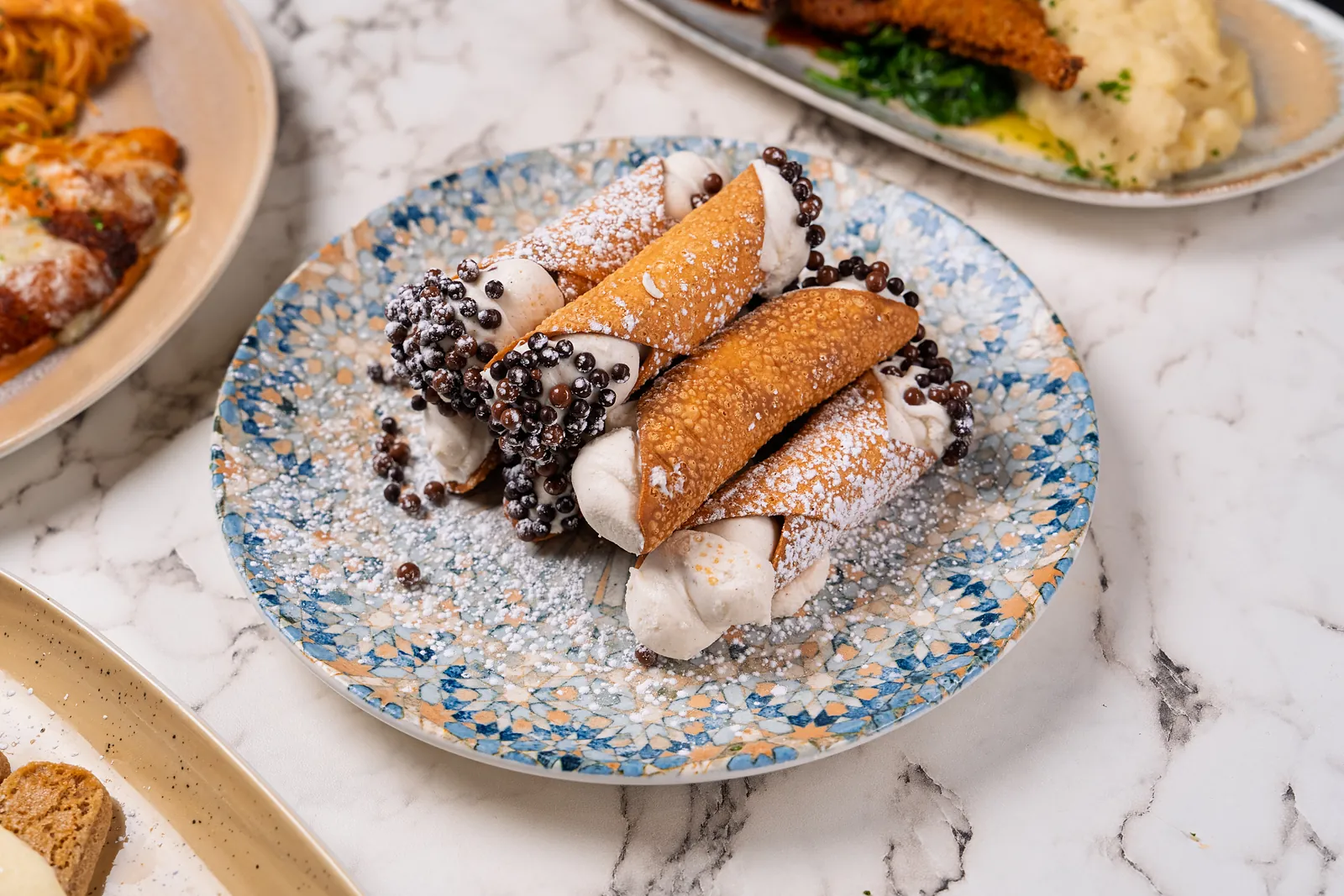 Three cannoli filled with cream filling and dipped in chocolate pearls on a blue speckled plate