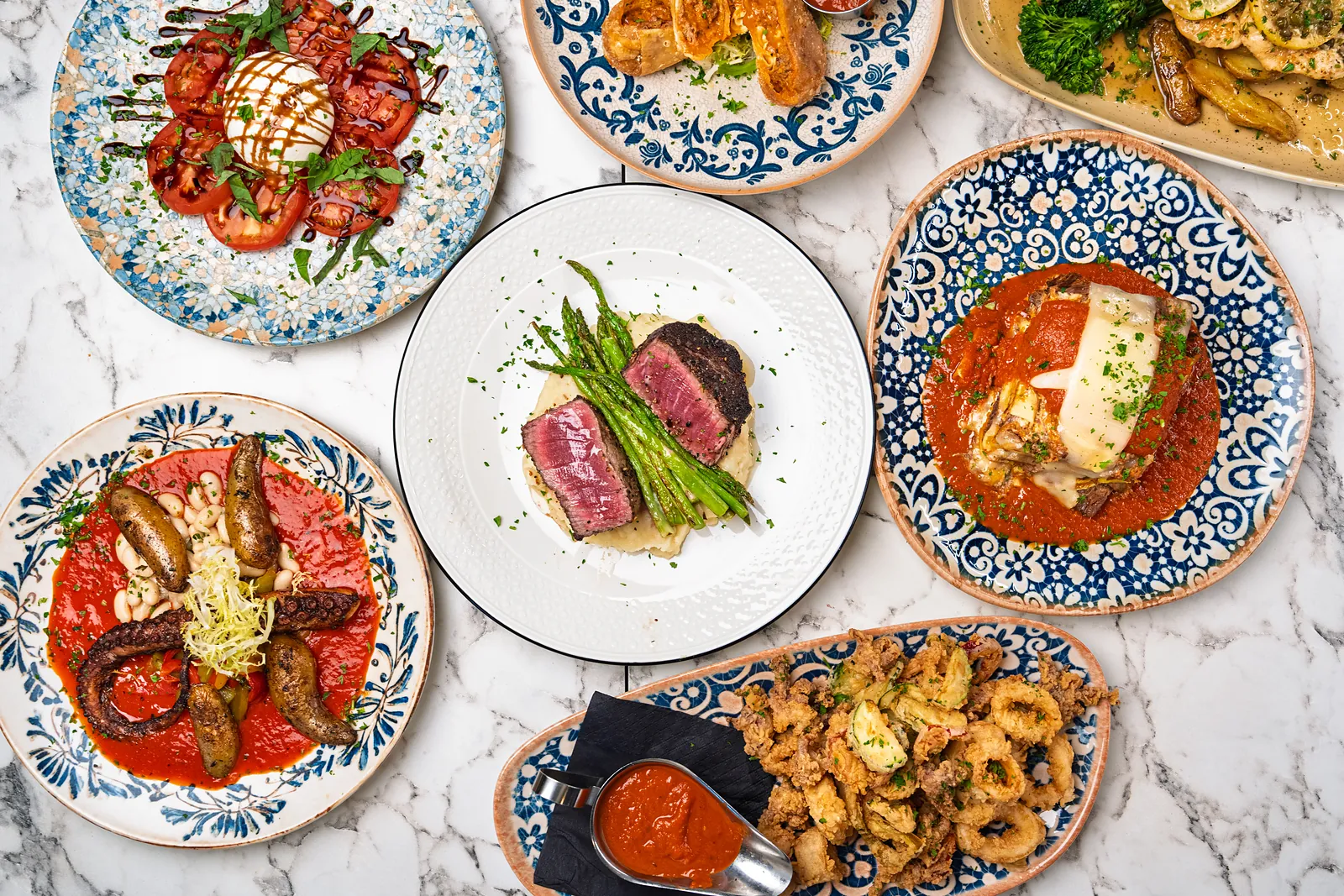 Overhead view of a marble table with filet and asparagus, burrata and tomatoes, grilled octopus, parmigiana and fried calamari on blue and white plates