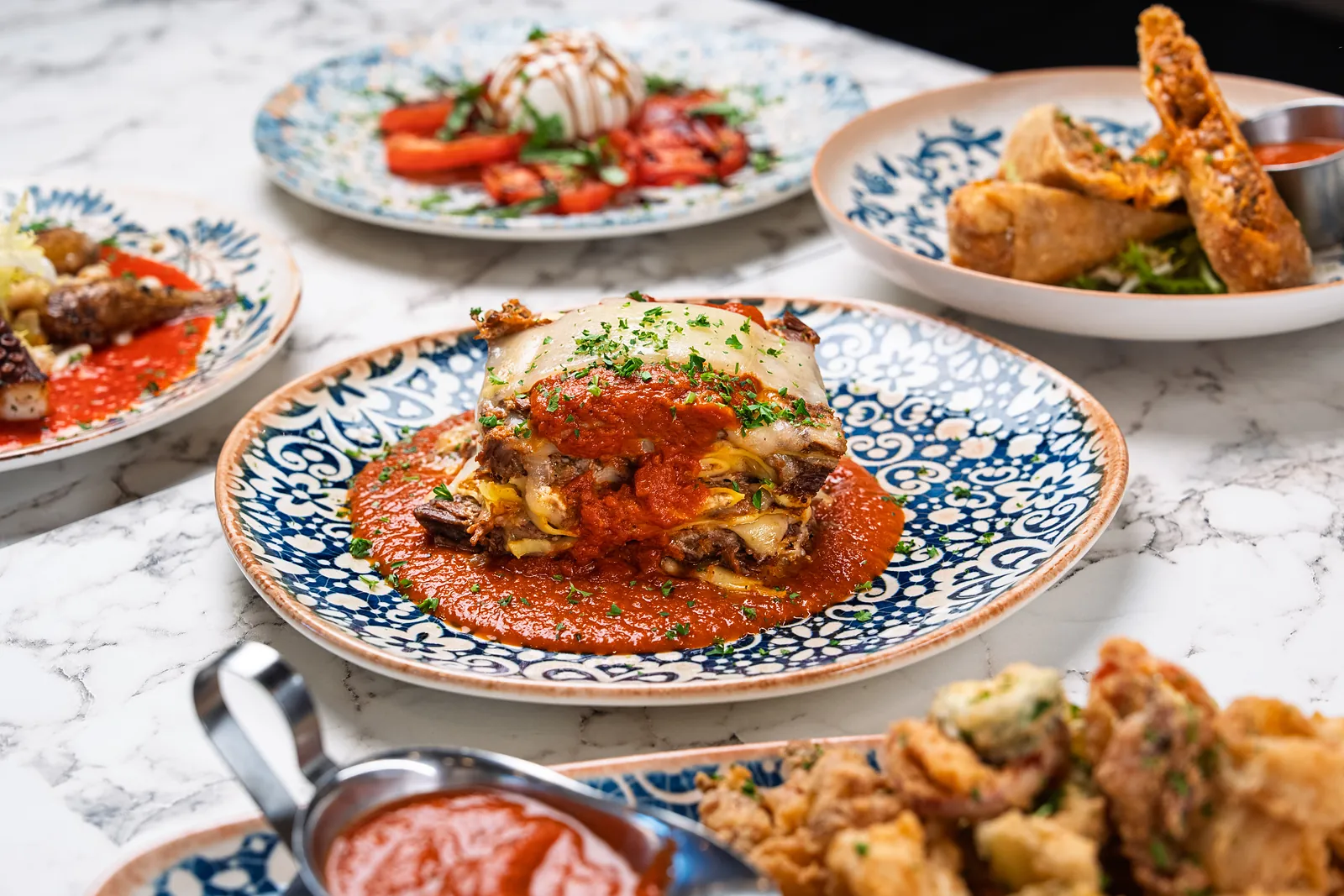 Table spread with a baked parmigiana in tomato sauce at the centre, burrata and tomato salad and fried rolls behind